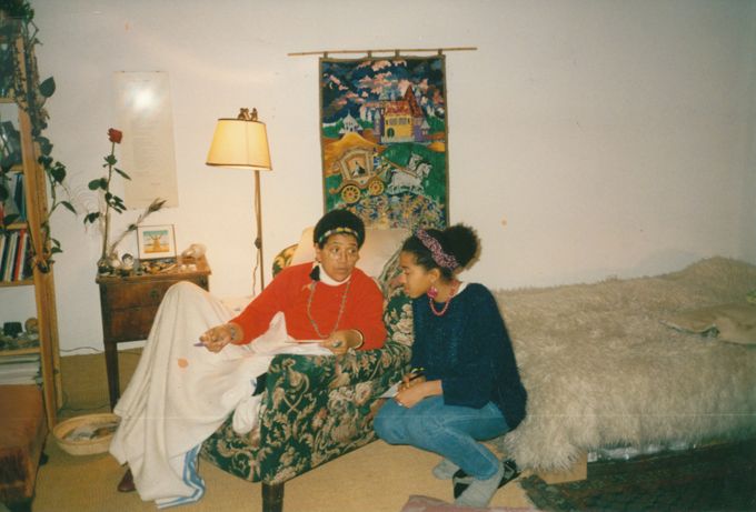 Audre Lorde (links) in einem Sessel sitzend und May Ayim (rechts) daneben kniend in einem Wohnraum; Bett und Regal im Hintergrund